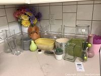 Variety of clear glass, ceramic, and metal floral vases and floral frogs arranged on counter.