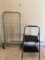 Metal shopping cart folded upright and two-step step ladder side by side on floor.