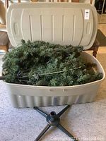 Gray plastic storage bin open to show artificial Christmas tree branches pre-strung with colored lights inside. Black tree stand base visible in front of bin.