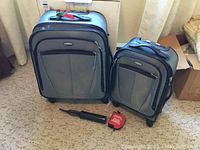 Two grey Samsonite Caribbea roller suitcases side by side, with a black manual air pump and a red survival kit pouch placed in front on carpeted floor.