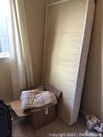 Twin boxspring mattress, beige quilt on a cardboard box, and off-white drapes hung on a window.