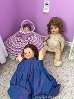 Set of three vintage dolls: purple crochet doll with bonnet, blue dress doll, floral dress doll sitting on carpet against purple wall.