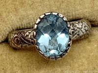 Close-up front view of the oval faceted blue stone set in a raised prong setting on the floral patterned sterling silver band.