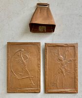 Front view of two copper fairy nude relief plaques alongside an antique copper cowbell, showing embossed cherub designs and overall condition.