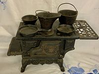 Front side view of the Crescent miniature cast iron stove with four miniature cookware pieces on stove top and side shelves extended. Stove features embossed Crescent logo and decorative legs.