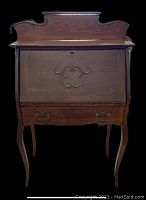 Front view of small antique oak drop front desk showing carved decorative front panel, drop front closed, curved legs, and ornate brass drawer pulls.