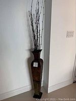Full view of the tall ceramic decorative floor vase placed on floor against light wall showing its size and shape with twigs inside.