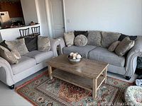 Photo showing the sofa and loveseat set arranged around a wooden coffee table on a patterned rug, with decorative cushions on the seating.
