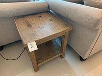 Wooden end table shown angled beside a sofa, featuring visible wood grain on top surface and lower shelf, with good condition and no visible damage.