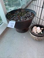 Two decorative flower pots placed on a balcony floor; one large dark pot containing soil, small plants, and black stones, the other smaller beige pot filled with shells and coral pieces.
