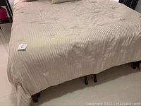 View showing queen sized bed with beige comforter, gently wrinkled, with some minor spots visible on fabric and black metal bed frame legs exposed below.