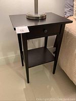 Full view of black wooden nightstand showing the drawer and lower shelf, placed next to a bed with lamp on top