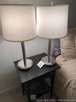 Pair of modern table lamps placed on a small black table between a bed and a wall, showing their white fabric drum shades and brushed metal cylindrical bases.