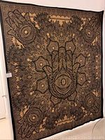 Photo showing full view of large cloth with detailed black Hamza hand design on beige background.