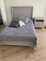 Full view of full sized bed made up with gray patterned bedding and assorted pillows, including white furry pillow. Bed frame is light wood with vertical slat headboard and solid footboard. Light wood two drawer nightstand with open shelf next to bed.