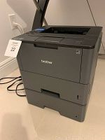 Front and side view of the Brother laser printer on floor near wall outlet, showing general shape and condition