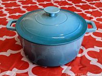 Blue enameled cast iron Dutch oven placed on a red and white patterned surface, showing front view with both handles and lid on.