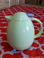 Front view showing the pastel green thermos pitcher with handle, rounded body, and blue feet and top sealing mechanism.