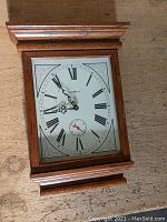 Front view of rectangular wooden wall clock with white face, Roman numerals, black ornate hands, and seconds subdial.