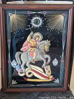 Full view of the framed textile icon showing Saint George on horseback slaying the dragon with metallic thread and bead details, behind glass in dark wood frame.