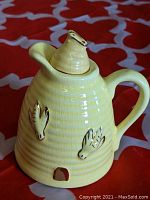 Front side view of yellow glazed ceramic beehive honey pot with gilt bee details and original lid on red and white patterned surface.