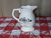 Front side view showing the printed logo and text on the white porcelain pitcher with handle and scalloped spout.