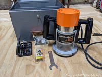 Photo showing the router with orange top and black handles, included router bits, metal wrench, and steel carrying case behind.