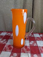 Front profile of tall orange and white cased glass pitcher with clear handle on right side and spout at top.