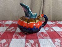 Front angle view of an art pottery pitcher shaped like a gourd with multicolor glazes including red, brown, blue, and green on a fabric surface.