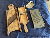 Image showing the three antique kitchen tools lying on a blue cloth background: large wooden cabbage slicer, two-sided wooden slicer/julienne, and metal Magic grater.