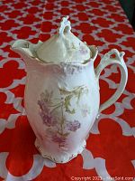 Front angle of cream-colored porcelain chocolate pot showing floral decoration and ornate lid.
