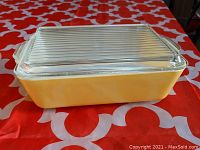 Front view of rectangular yellow Pyrex refrigerator dish with clear ribbed glass lid on orange and white patterned tablecloth.