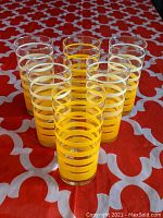Front view of seven clear tall glass tumblers with yellow horizontal bands set on red patterned surface.