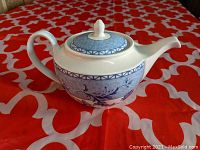 Side view of white ceramic Wedgwood teapot with blue Mikado floral and geometric pattern on a red and white fabric surface.