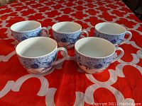 Five white ceramic cups with blue floral Mikado pattern arranged on red and white patterned cloth, photo taken to show front and side views of the cups.