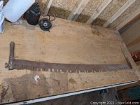 Full length photo of the large two-man crosscut saw laid flat on a workbench, showing wooden handles on either end and the long steel blade with perforated lance tooth pattern.