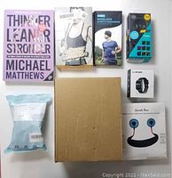 Top layout photo showing all boxed items arranged on a white surface including book, headphones, skipping rope, resistance bands, smartwatch, neck fan, and shakers.