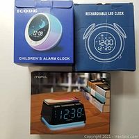 Boxes of ICODE Children's Alarm Clock and rechargeable LED clock showing front and top designs