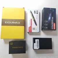 Top view of five boxed items including yellow EIOUMAX safe book, pen camera box, Korklian K68 anti spy detector box, wristwatch camera box, and clock camera box.