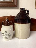 Two antique vessels side by side on wooden surface against white wall: one tall ceramic crock with brown glazed top, unglazed bottom with handle; smaller white glass vessel with Glenmore whiskey logo and cork stopper
