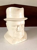 Angle showing the face side of the cream ceramic mug shaped as Winston Churchill's head wearing a hat and bowtie.
