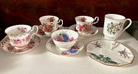 Front view of five bone china cups and saucers in floral patterns with gold trim arranged on white surface.