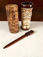 Full view of two tiki ceramic cups and a wooden tiki letter opener on white surface with dark wood background.