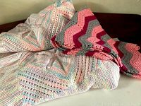 Two knit afghans displayed on a dark surface. One afghan is pastel multicolored with striped and open knit patterns, the other is chevron patterned in pink, red, and gray.