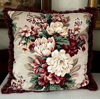 Front view of large square pillow showing the floral barkcloth pattern with cream and burgundy flowers and green leaves; moss fringe visible around edges.