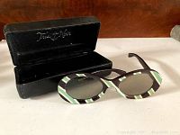 Opened black protective case displaying the sunglasses inside. The case has a velvet interior with silver 'Tous des Nil' inscription.