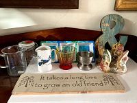 Wooden sign with friend quote, three fish figurines, blue ampersand, metal goblet, four small blue and white plates, plus pitchers and glassware arranged on wood shelf.