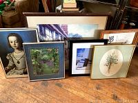 Six framed art pieces grouped on wooden floor showing variety in subject matter and frame styles