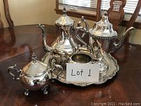 Full view of silver tea set on wooden table showing all five pieces arranged on serving tray with a lot label in front.