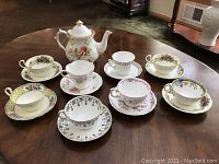 Eight assorted fine china cups and saucers displayed with a matching tea pot on a wooden surface.
